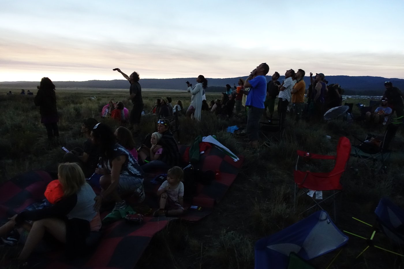 A crowd of eclipse watchers gathered outdoors at dusk, looking up at the sky